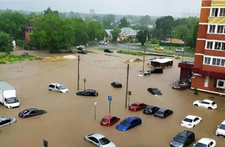 Mașini de diferite culori sunt parcate sau circulă pe un drum acoperit de apă de culoare maro, lângă clădiri și copaci sub un cer înnorat.