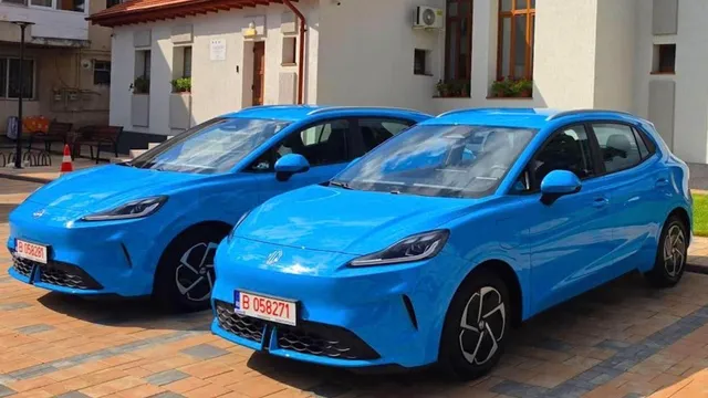 Două autoturisme electrice de culoare albastră sunt parcate pe o suprafață pavată în fața unei clădiri albe.