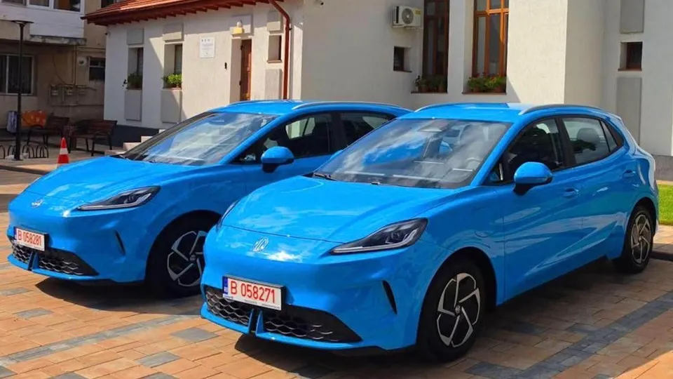 Două autoturisme electrice de culoare albastră sunt parcate pe o suprafață pavată în fața unei clădiri albe.