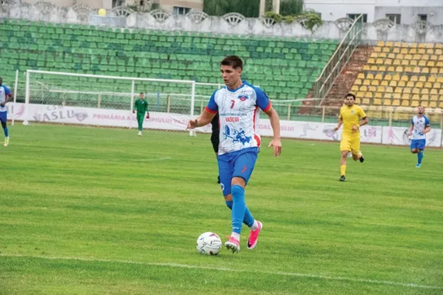 Un jucător de fotbal într-un echipament albastru și roșu conduce un balon pe un teren de fotbal verde în fața unor tribune goale.