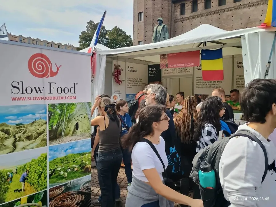 Oamenzi stau în fața unui stand alb care prezintă imagini cu peisaje și un logo cu un melc roșu sub textul Slow Food.