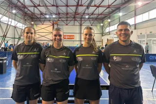 Patru persoane în echipament sportiv gri stau în rând într-o sală de sport cu mese de tenis de masă în fundal.