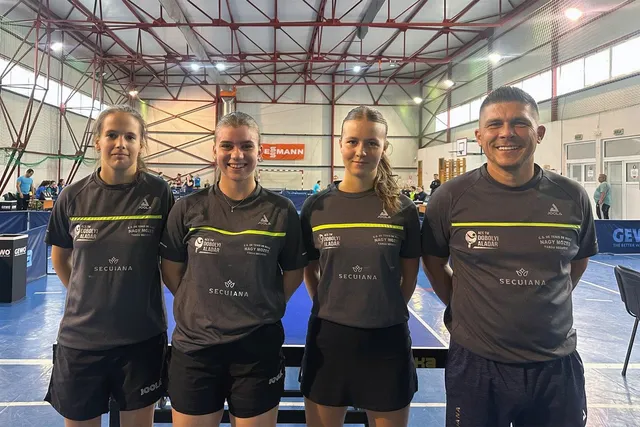 Patru persoane în echipament sportiv gri stau în rând într-o sală de sport cu mese de tenis de masă în fundal.