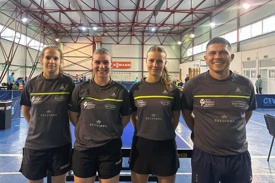 Patru persoane în echipament sportiv gri stau în rând într-o sală de sport cu mese de tenis de masă în fundal.