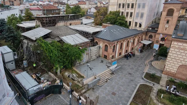 O vedere aeriană prezintă clădiri vechi cu acoperișuri metalice și de țiglă, un pavaj pietruit și câteva persoane pe un spațiu deschis.