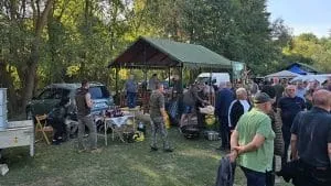 Un grup de oameni stă pe un teren verde lângă un cort mare și câteva mese cu obiecte de gătit în aer liber.