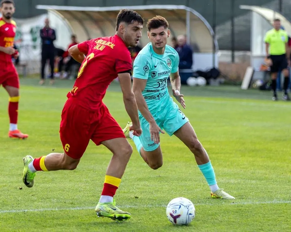 Un jucător în echipament roșu aleargă lângă un jucător în echipament turcoaz care conduce un fotbal pe un teren de fotbal verde.