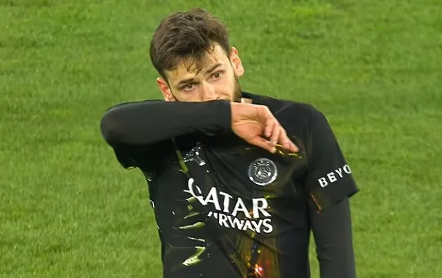 Un bărbat cu părul scurt și barbă poartă un tricou negru cu inscripții albe pe un teren de iarbă verde.