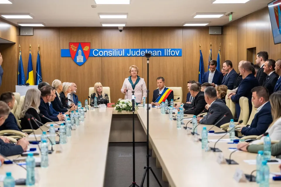 O grupă de persoane stă la mese lungi de conferință într-o sală cu pereți din lemn, în fața unui panou albastru cu inscripții.