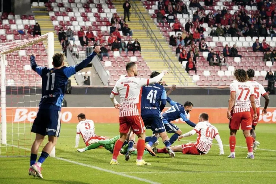 Jucători în tricouri albastre și roșii cu dungi albe, unul cu brațele ridicate în fața unei poți de fotbal, pe un teren verde cu spectatori în tribună.