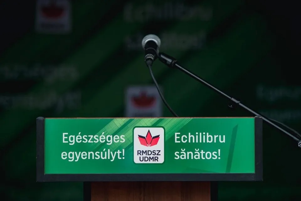 Un microfon negru este poziționat deasupra unui panou dreptunghiular verde pe care scrie text în limba maghiară și română.