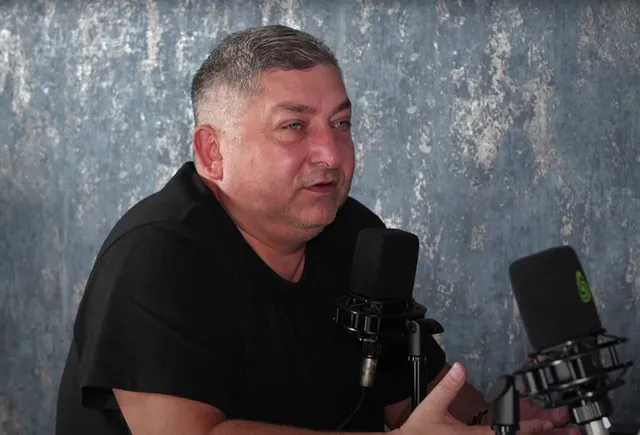 Un bărbat cu părul scurt și cămașă neagră stă în fața a două microfoane de studio pe un fundal gri texturat.