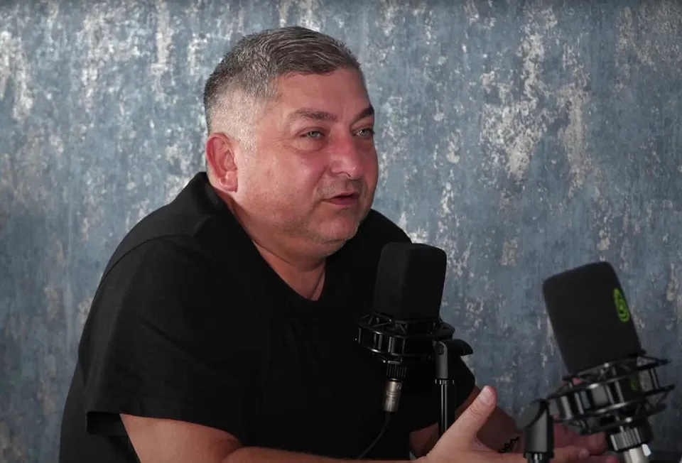 Un bărbat cu părul scurt și cămașă neagră stă în fața a două microfoane de studio pe un fundal gri texturat.