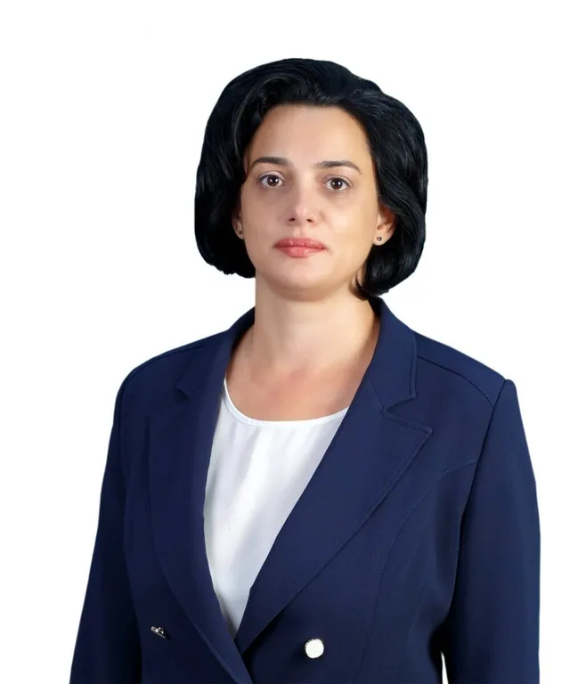 O femeie cu părul negru, purtând un sacou albastru peste un tricou alb, privește spre cameră pe un fundal alb.