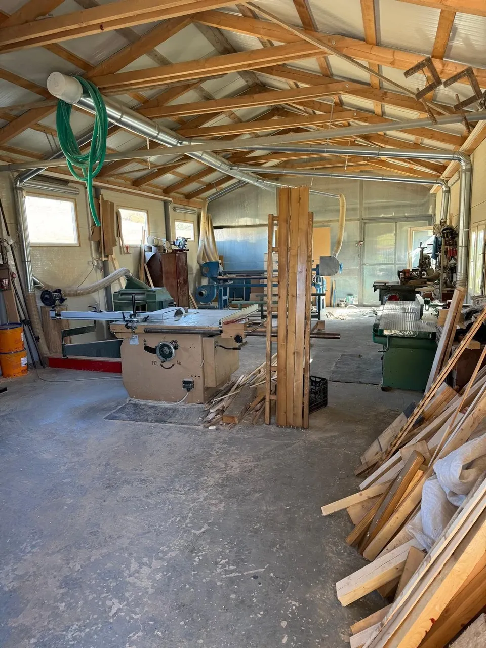 Un atelier cu grinzi de lemn expuse conține utilaje de lucru, materiale de construcție și un furtun verde suspendat de tavan.