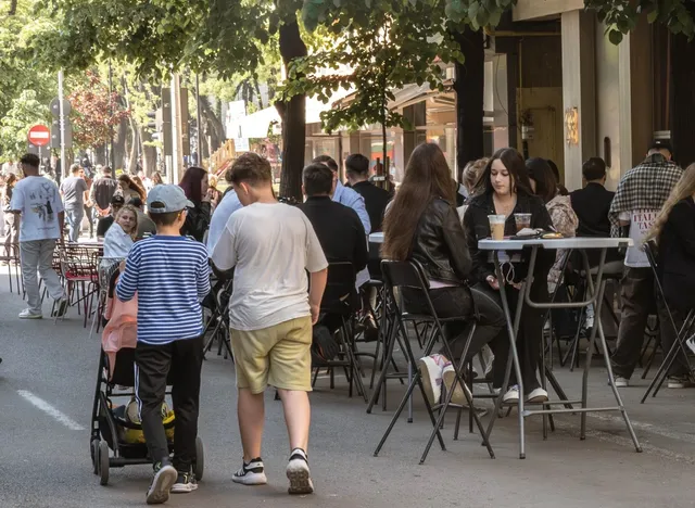 Oamenii stau la mesele de afară ale unor cafenele de pe un bulevard cu copaci și pietoni care merg pe trotuar.