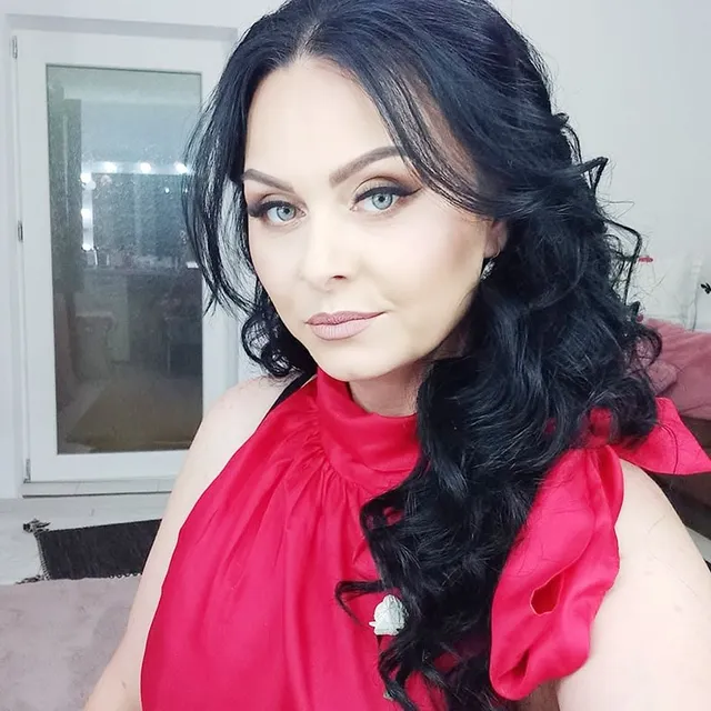 O femeie cu părul negru și lung, purtând o bluză roz, privește spre cameră în fața unei ferestre albe.