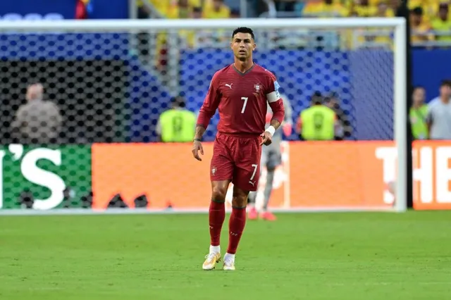 Un fotbalist într-un echipament roșu cu numărul șapte stă pe un teren de fotbal verde în fața unei porți de fotbal.