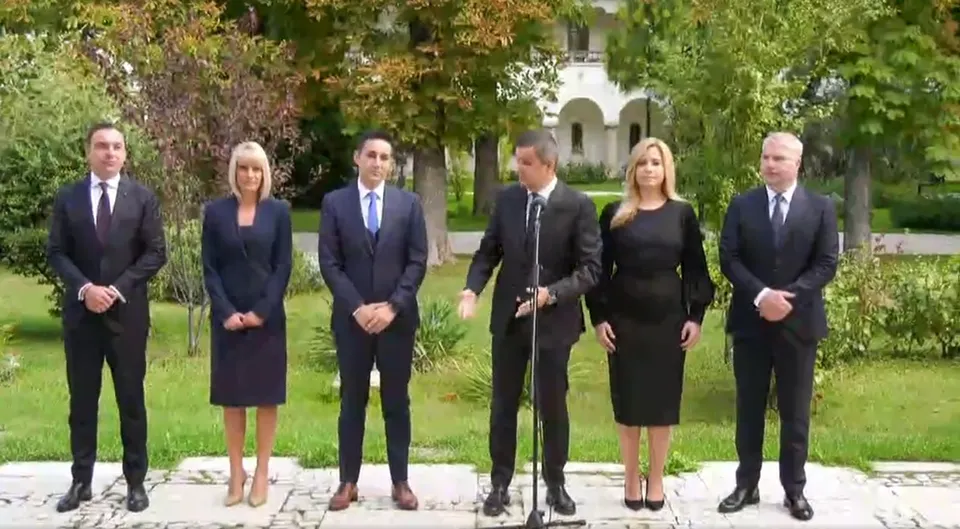 Șase persoane îmbrăcate în haine oficiale negre stau în rând pe un teren verde în fața unor copaci.