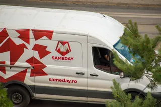 Un van alb cu logo-ul roșu Sameday și textul sameday.ro circulă pe un drum lângă ramuri de conifere verzi.