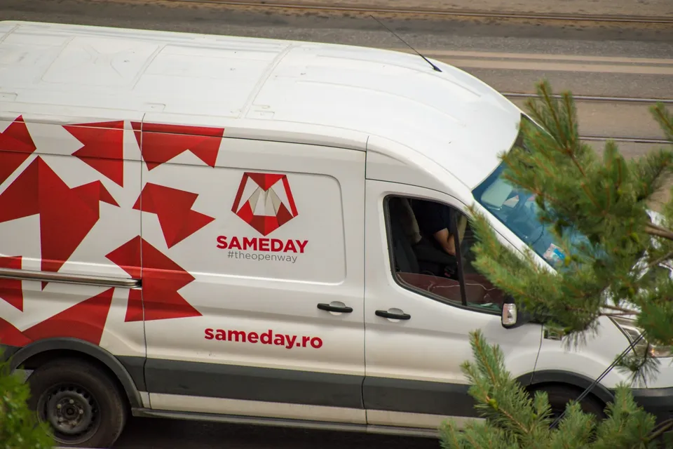 Un van alb cu logo-ul roșu Sameday și textul sameday.ro circulă pe un drum lângă ramuri de conifere verzi.