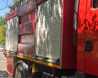 Partea laterală a unui camion roșu de pompieri prezintă compartimente cu capace metalice gri și inscripția POMPIERII scrisă cu litere albe.