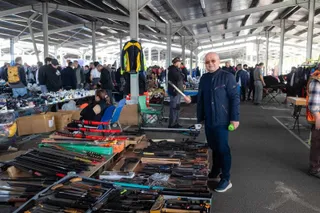 Un bărbat cu ochelari și o jachetă albastră stă în picioare lângă mese cu obiecte diverse într-un spațiu acoperit cu mulți oameni în fundal.