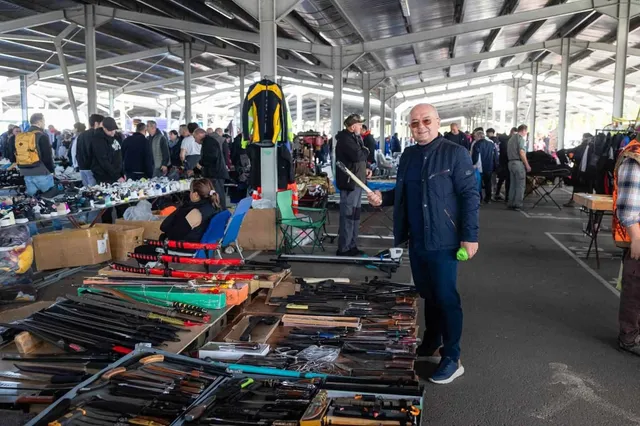 Un bărbat cu ochelari și o jachetă albastră stă în picioare lângă mese cu obiecte diverse într-un spațiu acoperit cu mulți oameni în fundal.