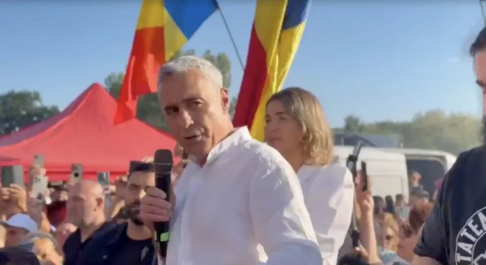 Un bărbat cu părul cărunt și cămașă albă vorbește într-un microfon în fața unei mulțimi și a unor steaguri colorate.