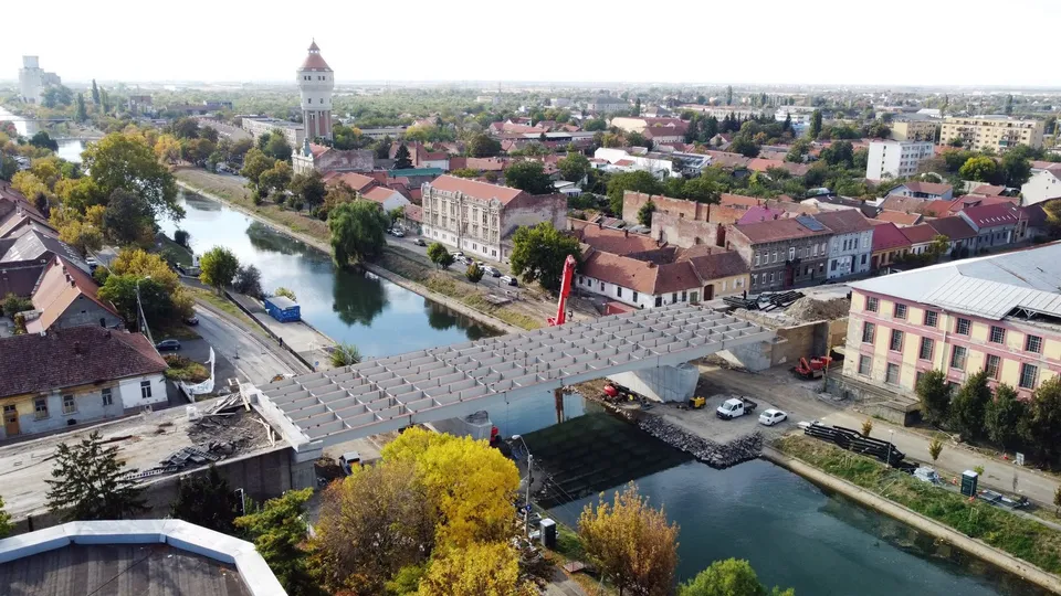 O vedere aeriană prezintă un pod metalic peste un canal care traversează o zonă urbană cu clădiri și copaci.