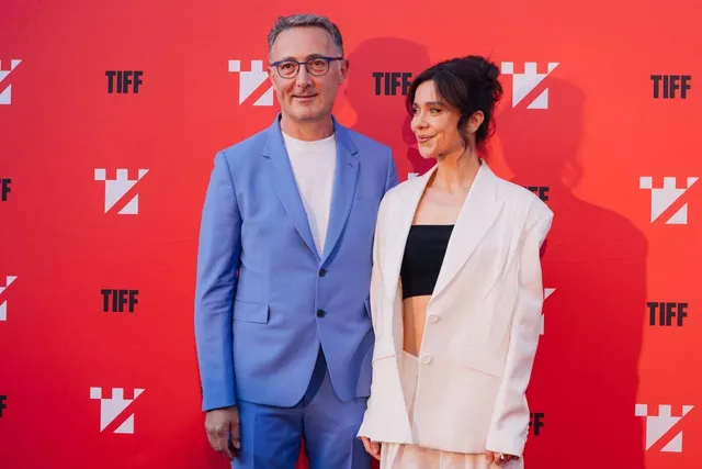 Doi actori, un bărbat și o femeie, stau în fața unui perete roșu cu logo-uri TIFF albe, la un eveniment.