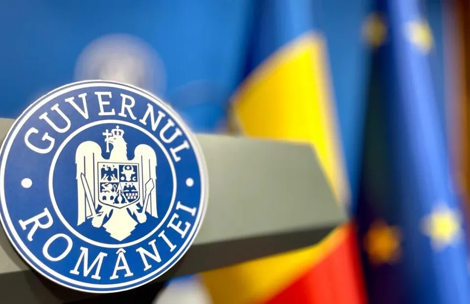 Un emblema circular albastru și alb cu textul Guvernul României este plasat în prim-plan, cu steaguri colorate în fundal.