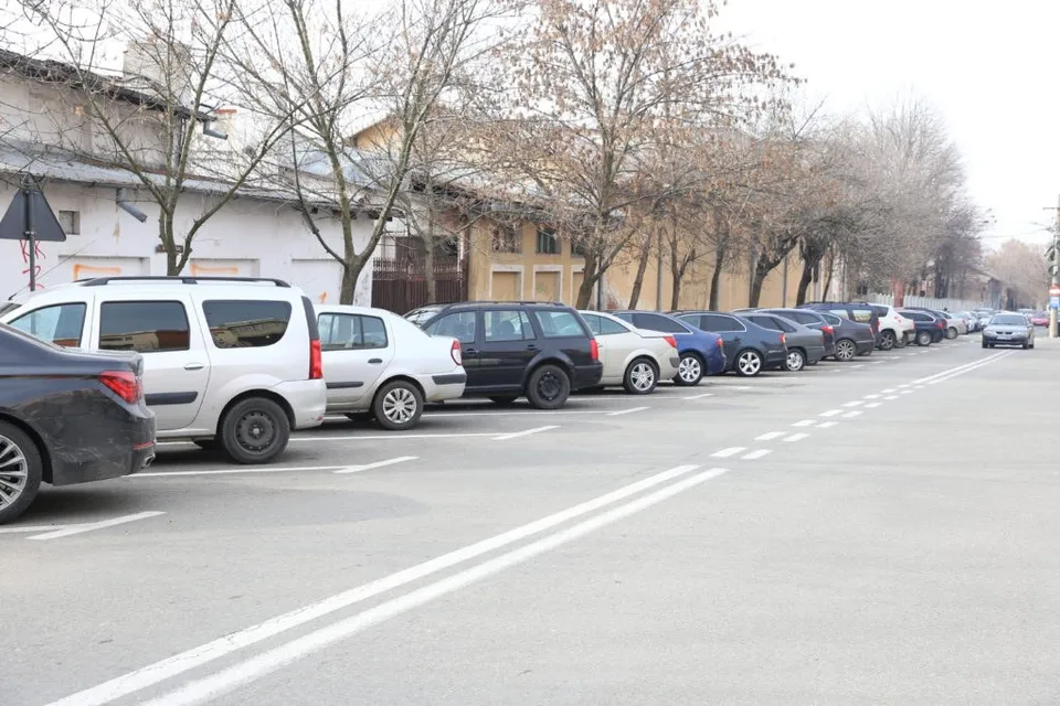 Mai multe mașini sunt parcate în rând pe un씩 loc de parcare marcat cu linii albe pe asfaltul unei străzi.