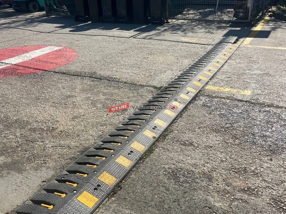 Un dispozitiv de tip bumper din plastic negru cu marcaje galbene este montat pe o suprafață asfaltată lângă o zonă marcată cu vopsea roșie și albă.