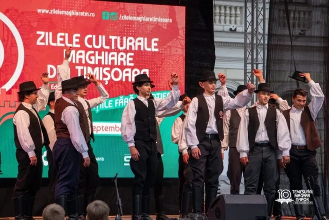 Un grup de bărbați îmbrăcați în costume tradiționale albe și negre execută un dans pe o scenă în fața unui panou roșu.