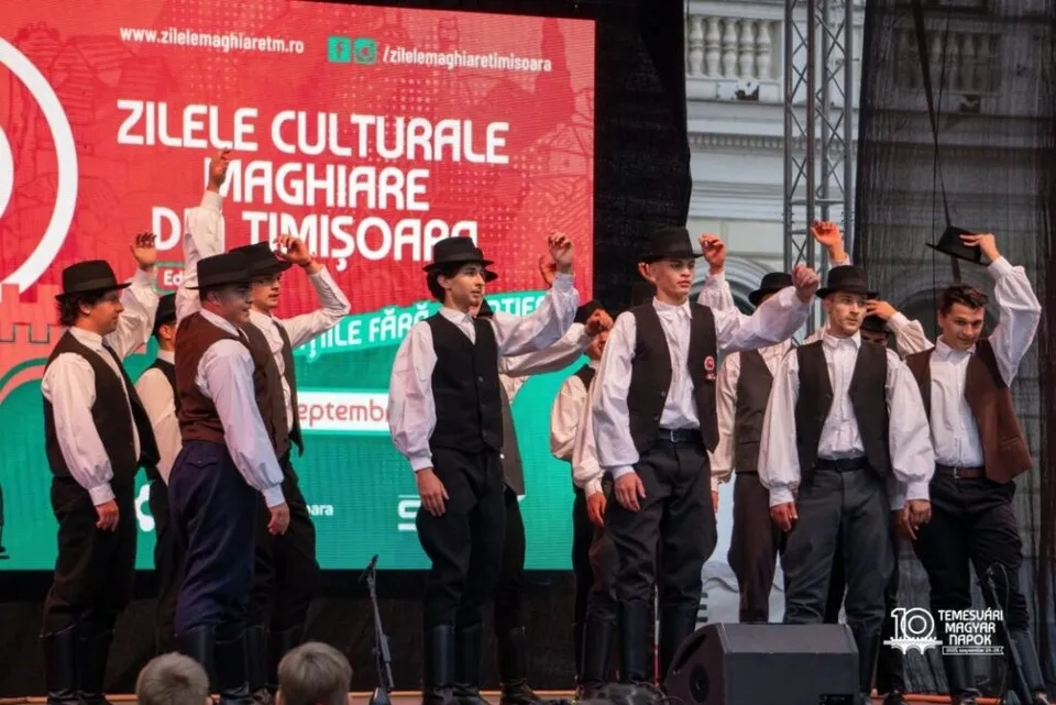 Un grup de bărbați îmbrăcați în costume tradiționale albe și negre execută un dans pe o scenă în fața unui panou roșu.