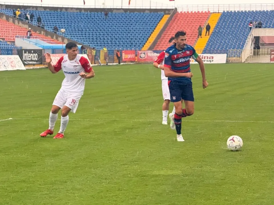 Un jucător de fotbal în echipament alb și roșu aleargă pe terenul de iarbă lângă un alt jucător în echipament albastru și roșu.