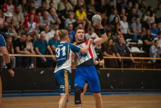 Un jucător de handbal în echipament alb și roșu ridică o minge, fiind interceptat de un adversar în echipament albastru și galben pe un teren.