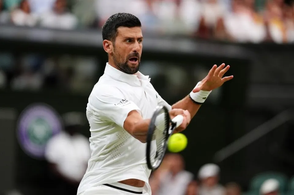 Un bărbat cu barbă, îmbrăcat în haine albe, lovește o minge de tenis galbenă cu o rachetă pe un teren de sport.