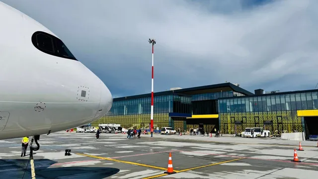 O parte din fuselajul unui avion alb este vizibilă în prim-plan, lângă un terminal de aeroport cu fațadă de sticlă și câteva vehicule pe pistă.