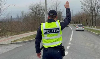 Un agent de poliție cu vestă reflectorizantă galbenă stă pe marginea unui drum și ridică mâna dreaptă spre o mașină argintie.