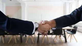 Două persoane purtând sacouri întemeiate își strâng mâinile într-un gest de strângere de mână într-o încăpere luminoasă cu scaune și mese.