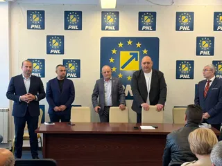 Cinci bărbați stau în picioare în fața unui perete decorat cu logo-uri albastre și galbene, în spatele unei mese de lemn întunecate.