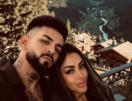 Un bărbat cu barbă și tatuaje pe gât stă lângă o femeie cu părul lung și machiaj pronunțat în fața unui peisaj montan cu case.