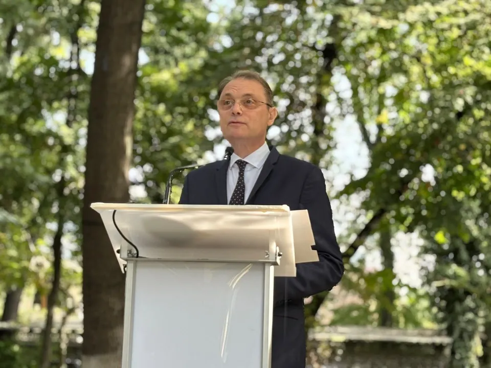 Un bărbat cu ochelari și costum închis la culoare stă în spatele unui podium alb într-un spațiu cu copaci verzi.