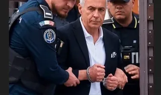 Un bărbat cu cămașă albă și sacou negru este condus de doi polițiști în uniformă întunecată printr-o ușă maro.
