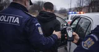 Un polițist în uniformă albastră ține de un bărbat cu hanorac negru lângă o mașină albă, în timp ce un alt agent folosește un aparat de testare.