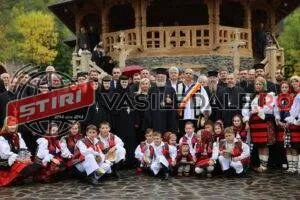 Un grup mare de oameni, inclusiv copii în costume tradiționale și adulți în haine negre, stau grupați în fața unei construcții din lemn.