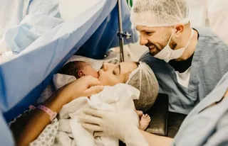 O femeie cu căciulă de protecție și un bărbat cu mască și căciulă de protecție stau lângă un nou-născut învelit în pânză albă.
