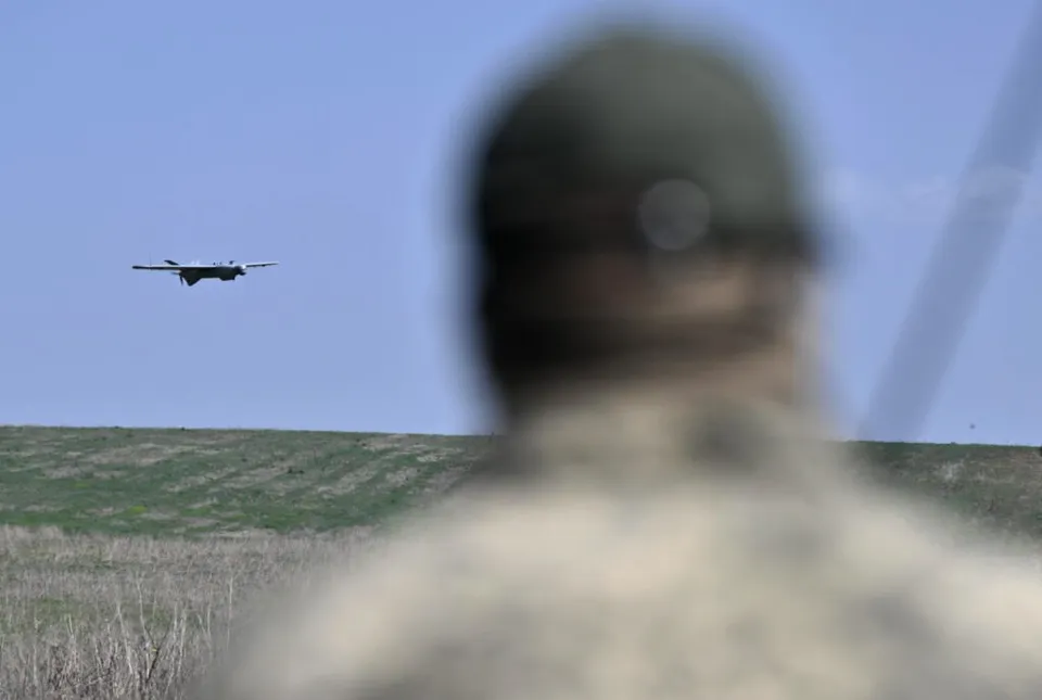 Un avion fără pilot zboară pe cerul senin deasupra unui câmp verde, în timp ce silueta neclară a unui soldat ocupă prim-planul.